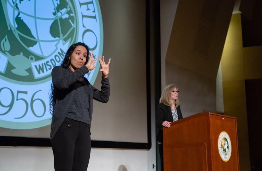 Woman offering ASL interpretation during speech, with USF seal behind her